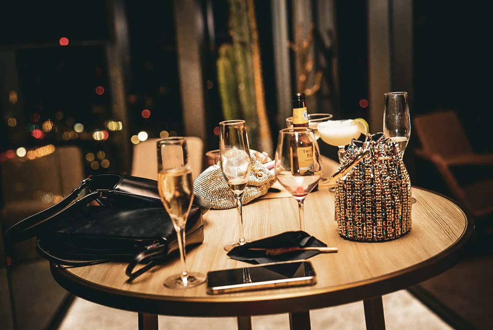 A round wooden table set with empty champagne glasses, a black clutch purse, a strappy black high heel shoe, a smartphone, a small clutch with gold accents, and a colorful, beaded bag. There are also two cocktail glasses with drinks, one with a lime 