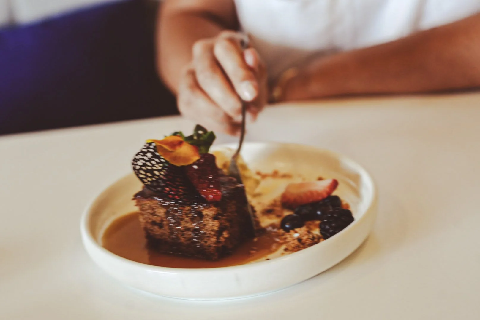 Close-up of a person using a fork to eat a slice of chocolate cake topped with berries and edible flowers, served on a white plate.