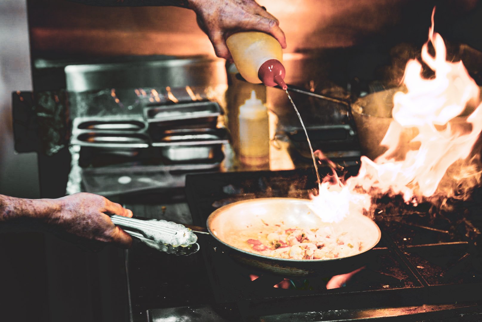 Chef pouring sauce into a frying pan with flames on a stove in a professional kitchen.