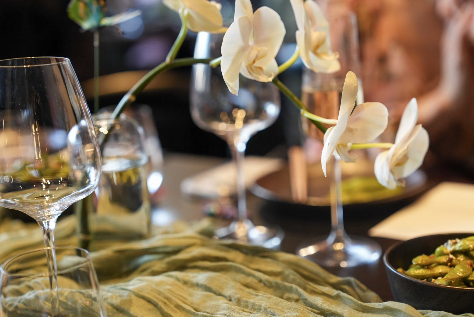 A table setting with white orchids in a glass vase, empty wine glasses, a bowl of vegetables, and a black dish, with a yellow fabric tablecloth.