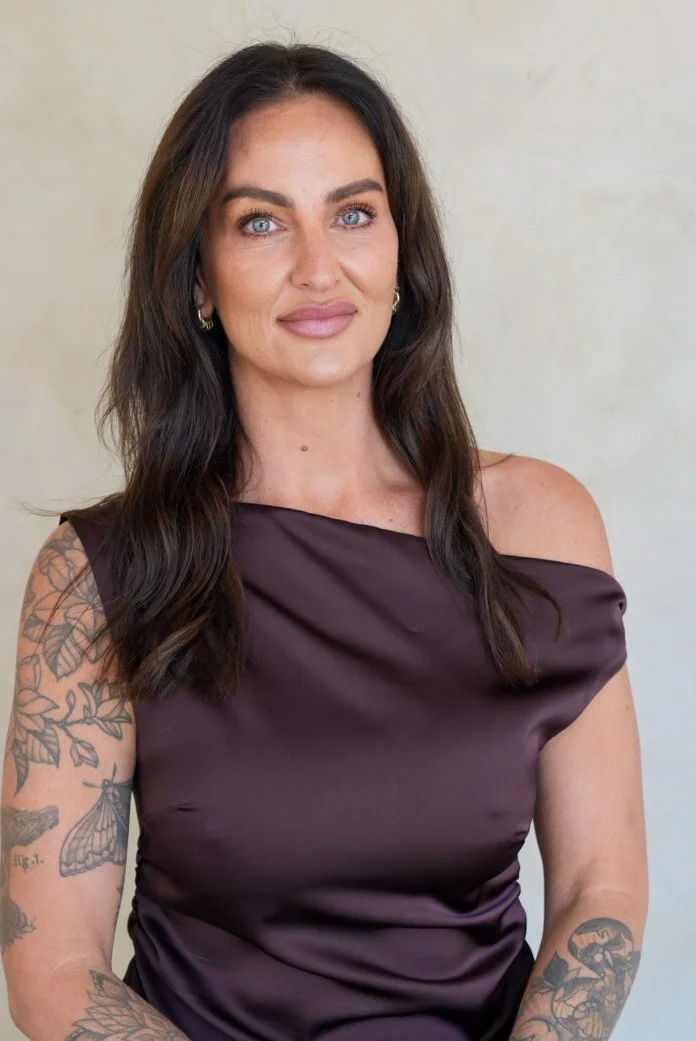 A woman with long dark hair, light blue eyes, and tattoos on her arm, wearing a dark purple off-shoulder top, standing against a plain light background.