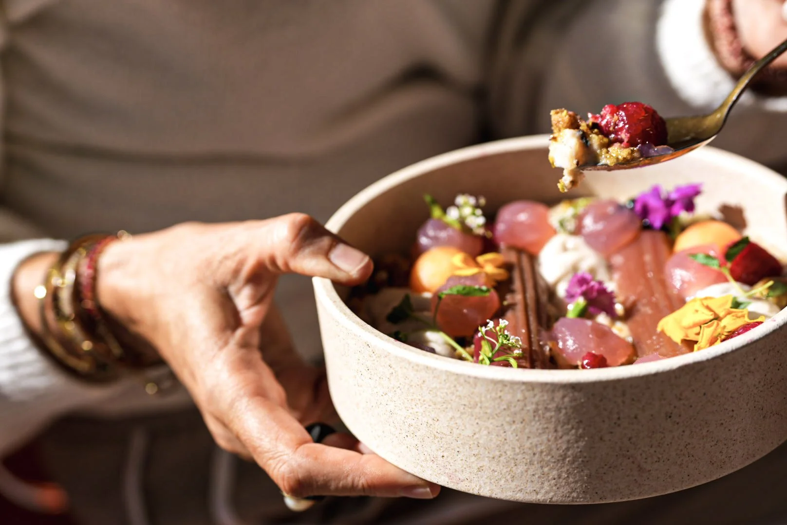 A person holding a bowl of colorful food, including pink and orange slices, garnished with edible flowers and herbs, using a spoon.