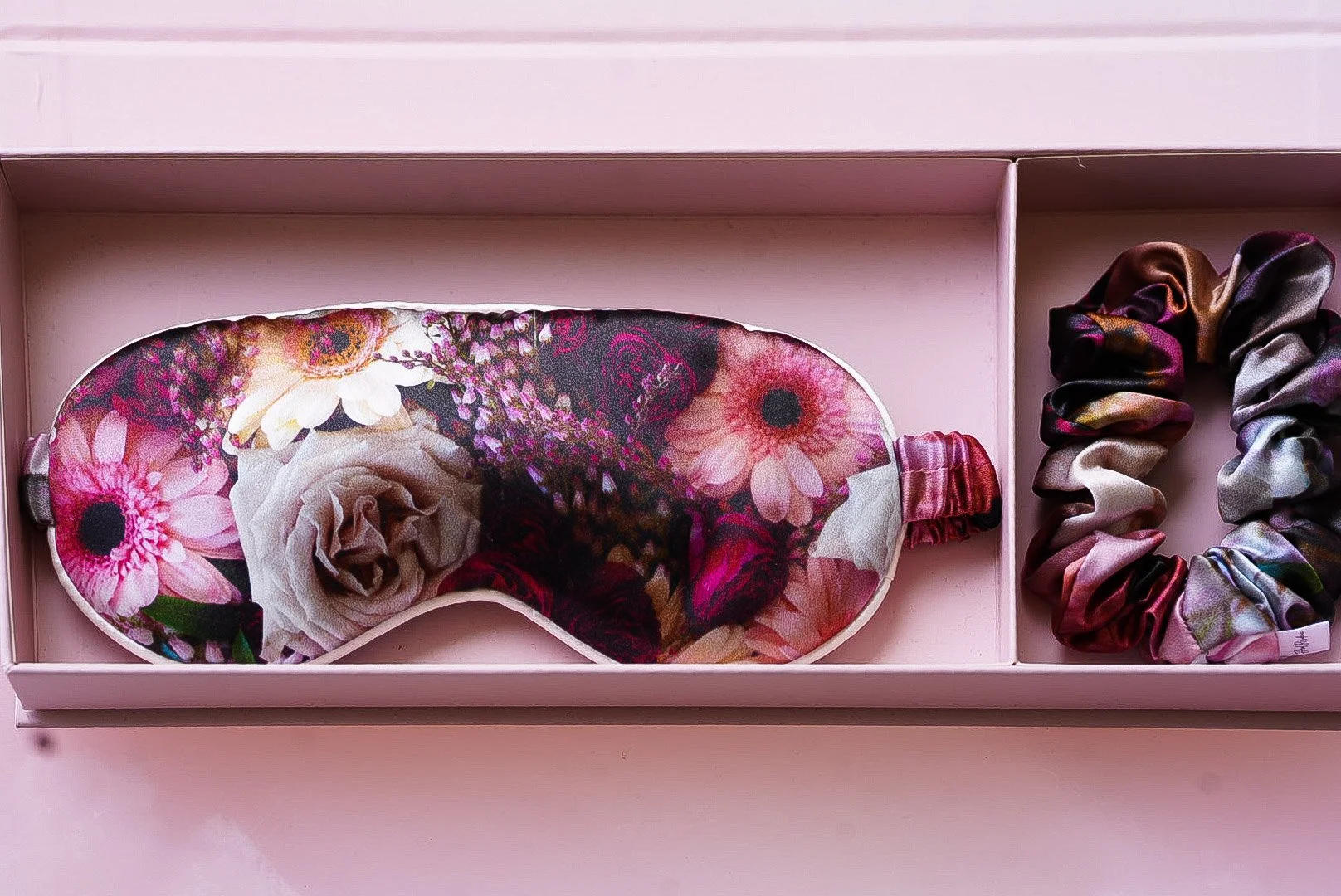 Pink box containing a floral sleep mask and a multicolored scrunchie.
