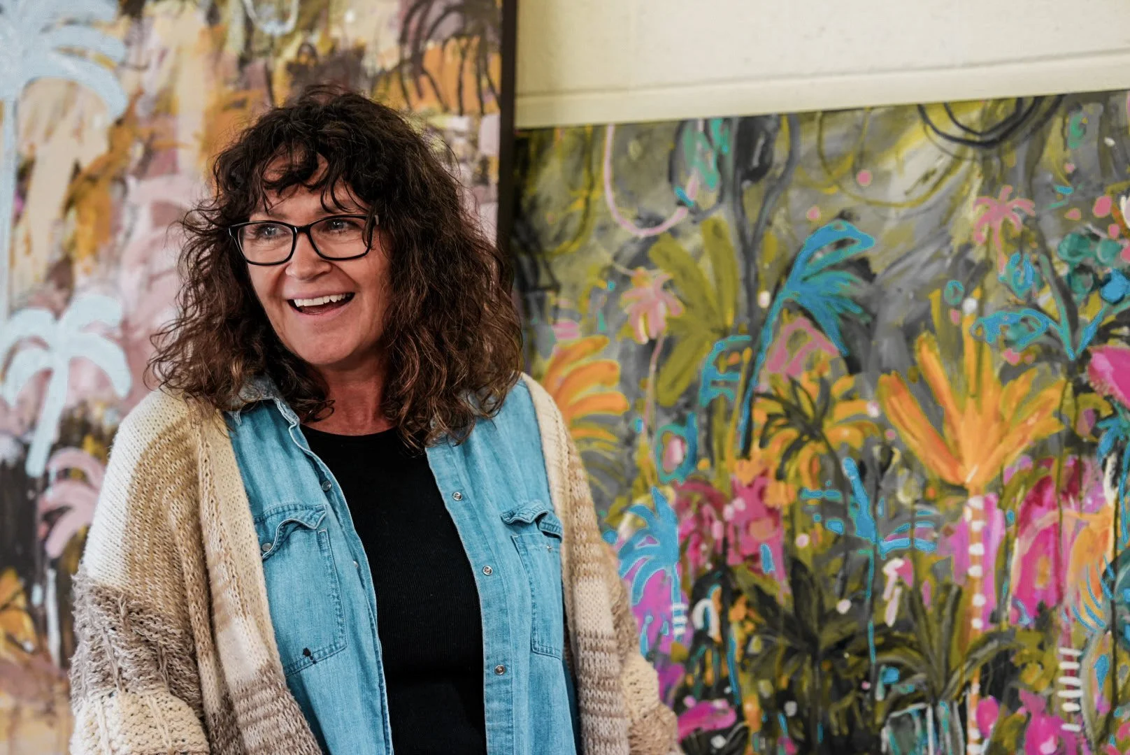 A woman with curly brown hair, glasses, wearing a denim shirt and a cream-colored knit cardigan, standing in front of colorful abstract floral paintings in an art gallery.