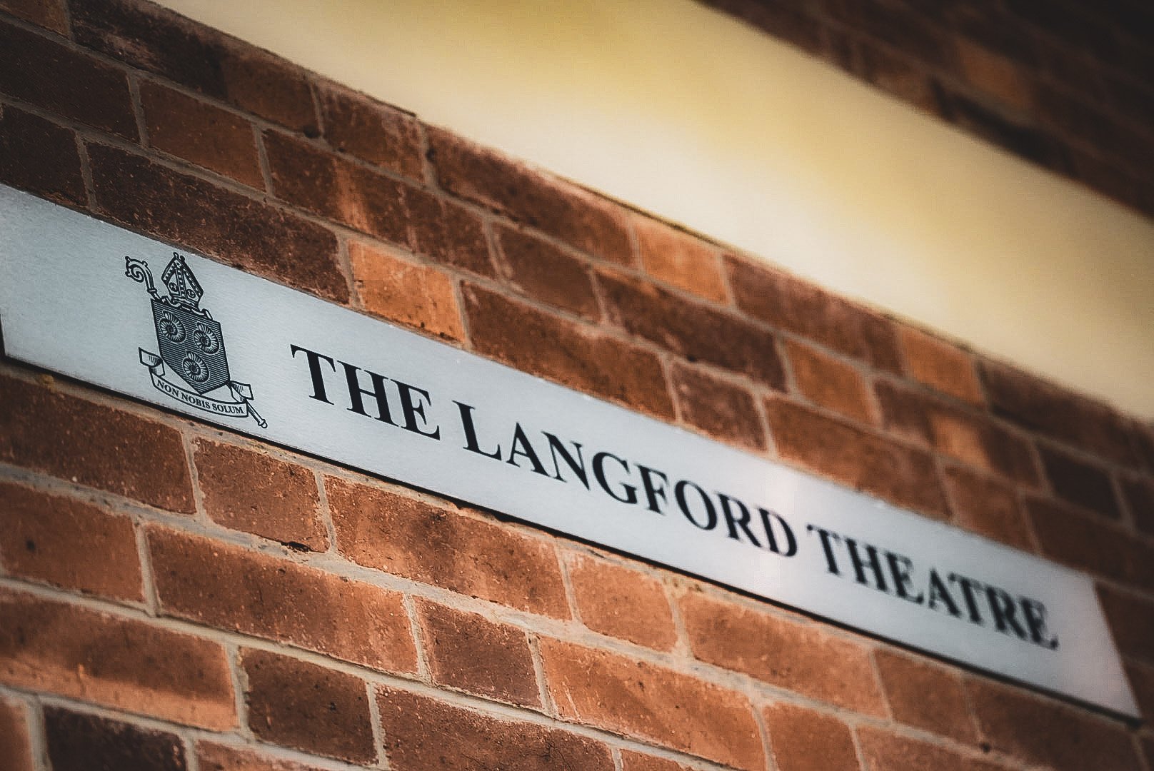 A brick wall with a white sign reading "The Langford Theatre" and an emblem featuring a shield with a crown on top and a banner below that reads "Non Nobis Solum."
