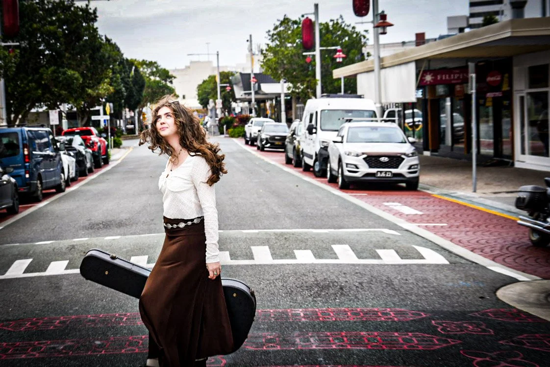 A young woman standing on a city street holding a guitar case, with parked cars and shops in the background.