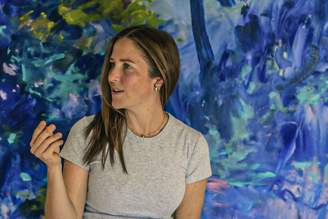 A woman with brown hair and freckles wearing a gray t-shirt, standing in front of a colorful abstract painting with blue and green hues.