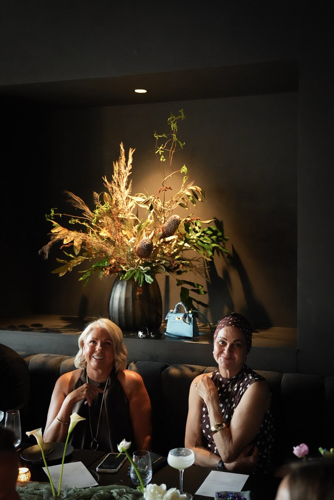 Two women sitting at a table in a dimly lit restaurant with a large floral arrangement in the background and a blue handbag on the shelf behind them.