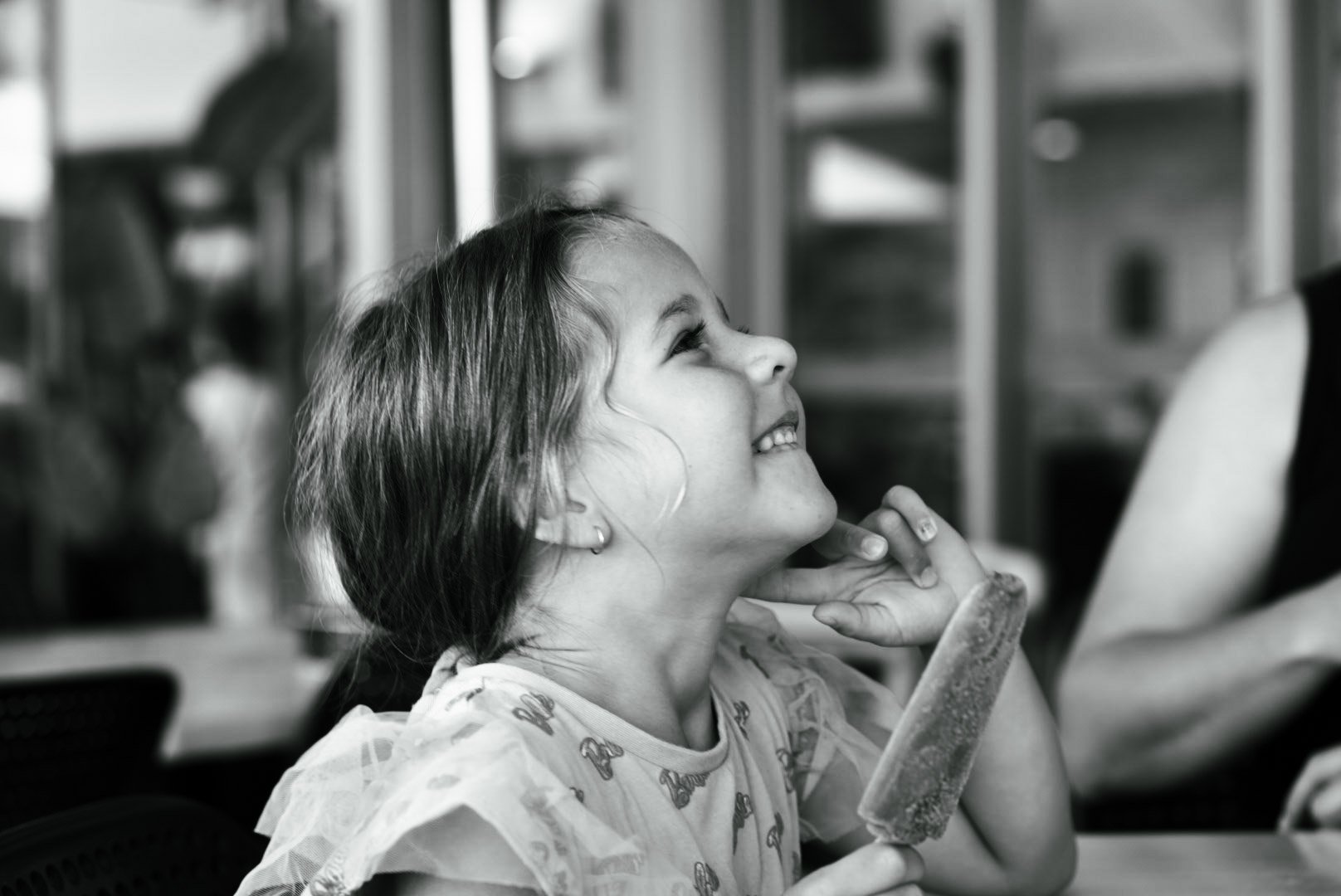 A young girl with a big smile, looking happily upward while sitting at a table with a partially eaten ice cream cone in her hand, in a black-and-white photo with a blurred background.
