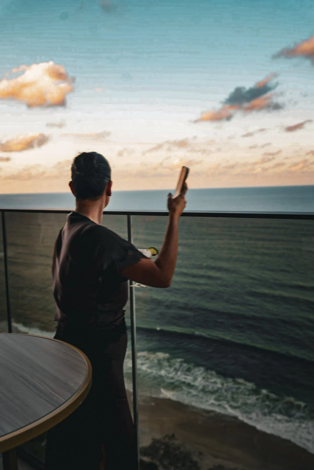 A person taking a photo of the ocean with a smartphone on a balcony during sunset.