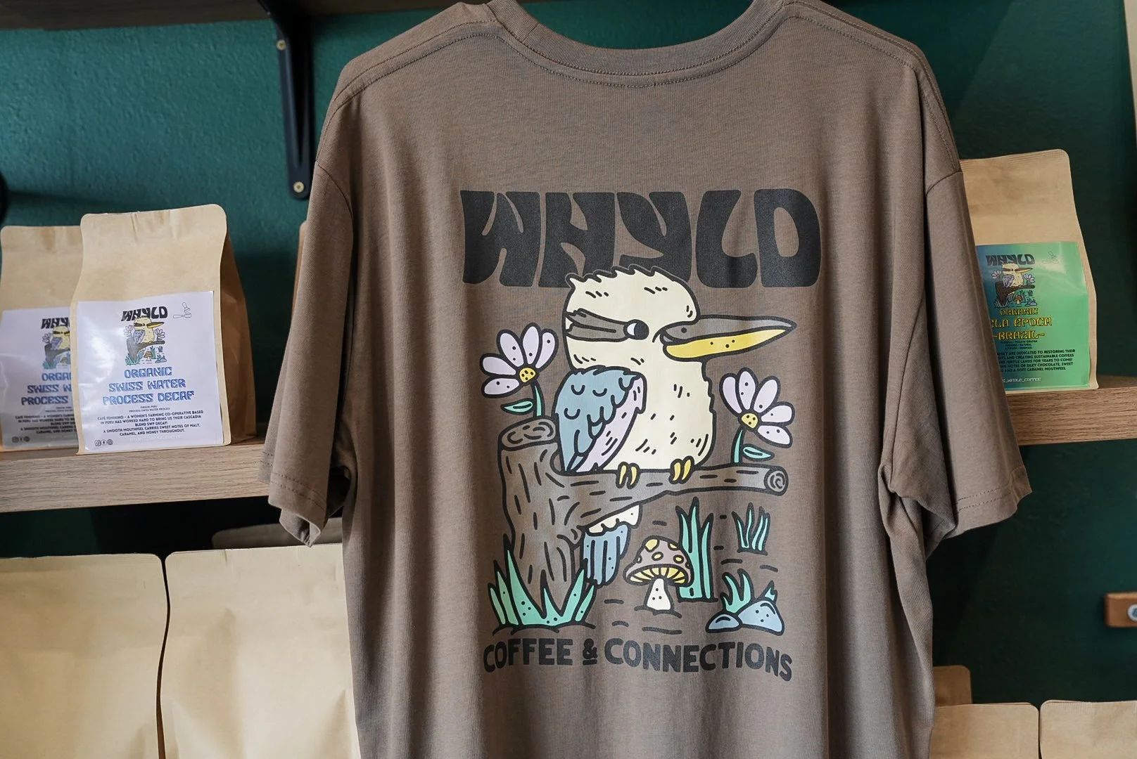 A brown T-shirt with a cartoon design of a pelican and a bird sitting on a branch, surrounded by flowers, mushrooms, grass, and rocks. The shirt has the text "WILD" above the design and "COFFEE & CONNECTIONS" below.