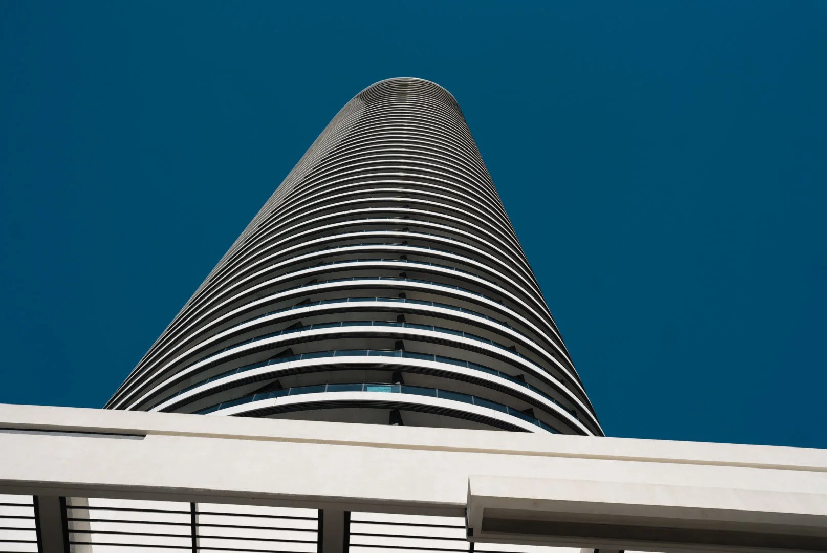Low-angle view of a tall, modern, curved skyscraper with horizontal white bands and balconies, against a clear blue sky.