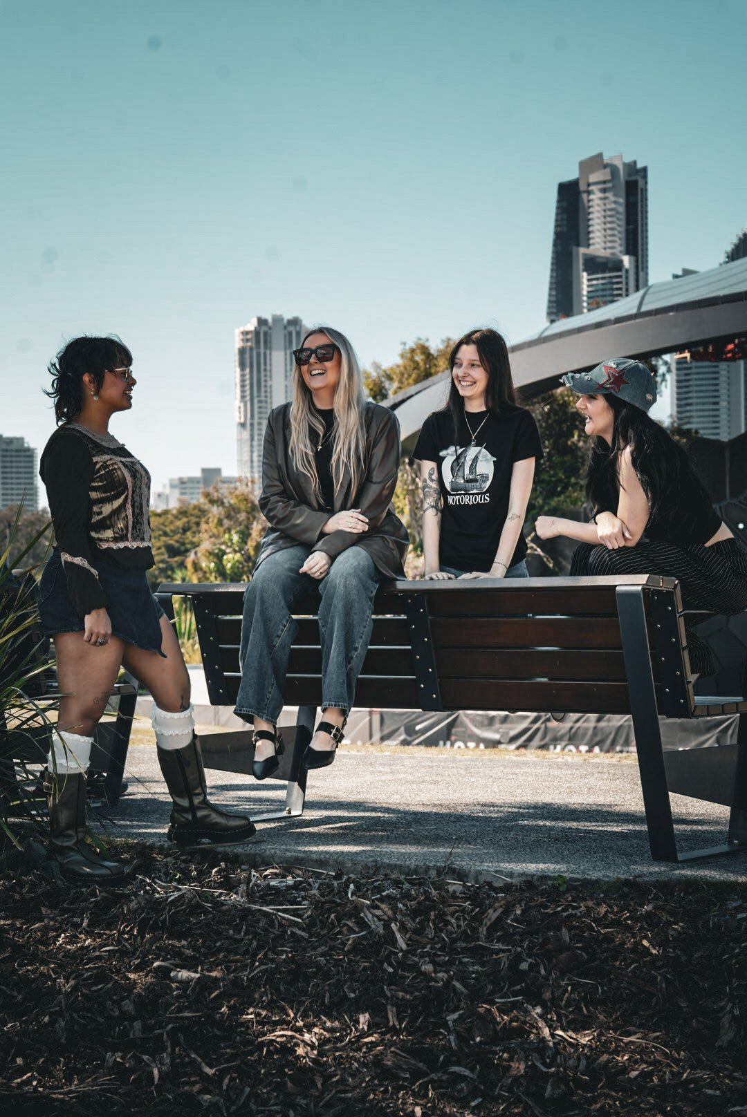 Four women of different ethnicities enjoying a conversation outdoors on a city street with tall buildings in the background. One woman with dark hair and glasses stands on the left, another with long blonde hair and sunglasses sits on a park bench, a