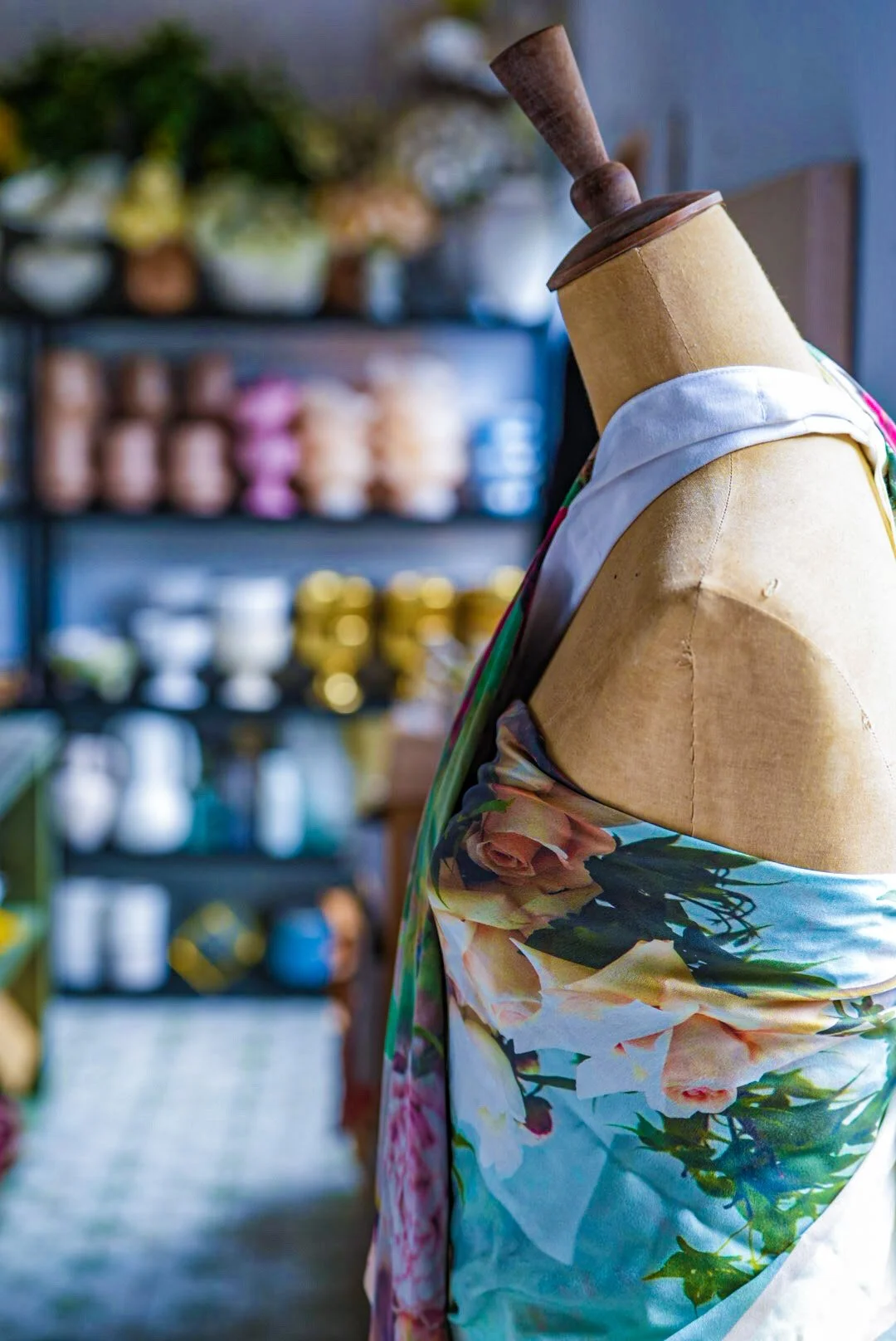 A mannequin on a stand wearing a floral patterned fabric or dress, with shelves of colorful ceramic pots and jars blurred in the background.