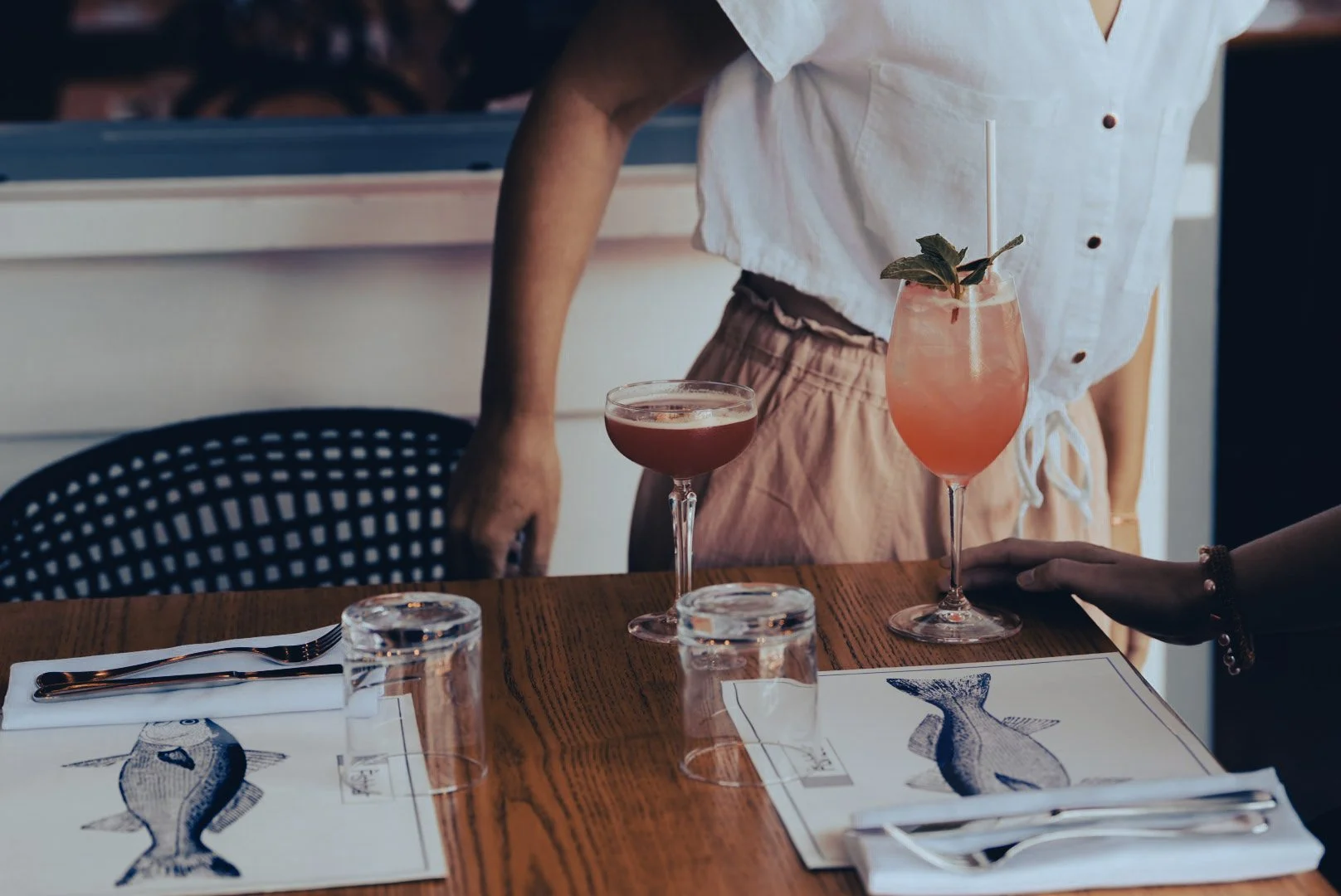 Two cocktails on a restaurant table, one pink drink decorated with mint and a straw, and a darker red drink in a coupe glass. Someone's hand is reaching towards the pink cocktail, and a person in a white shirt and beige pants is standing nearby.