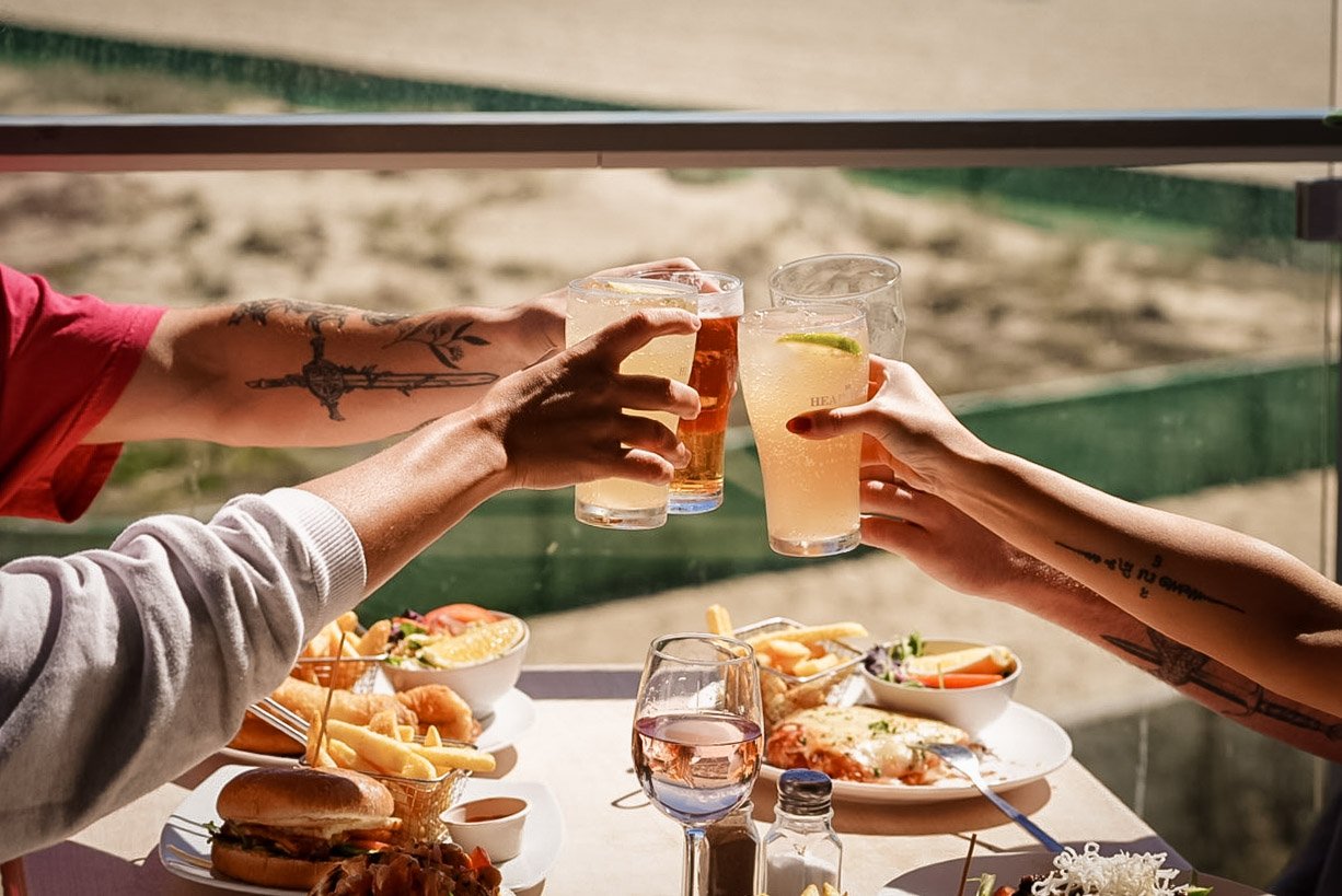 Three people clinking glasses of drinks with food on a table in front, outdoors, possibly at a restaurant or cafe by a beach or water.