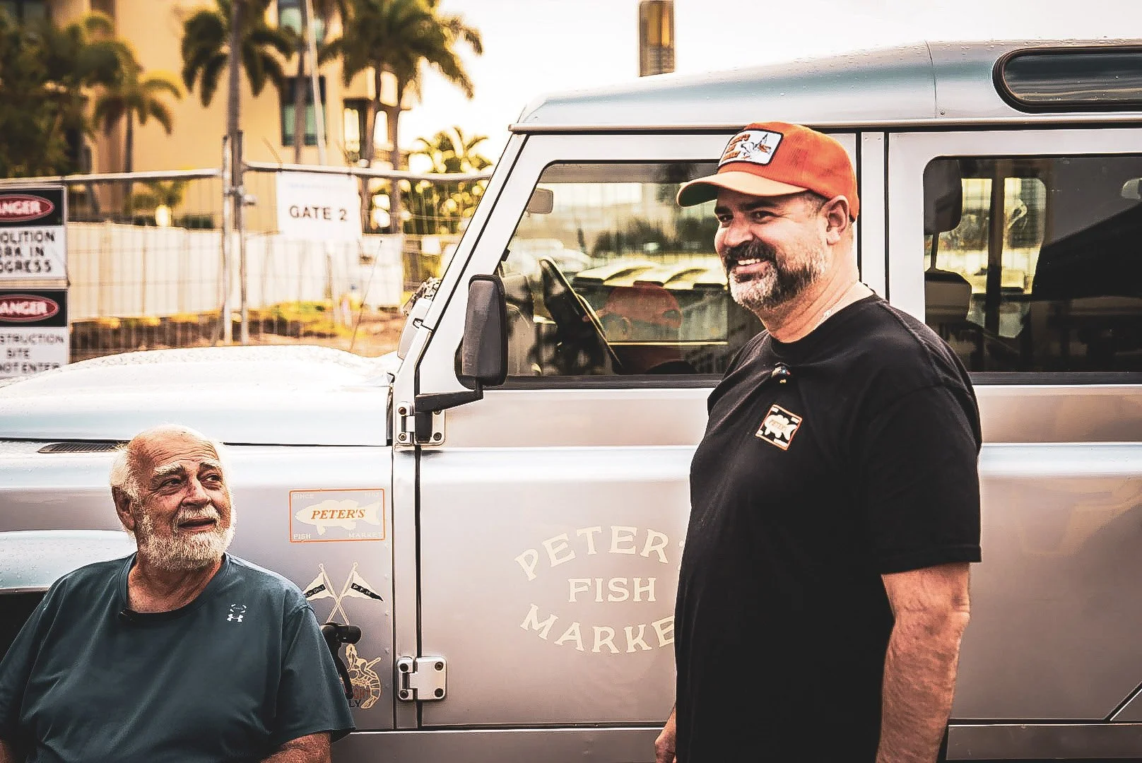 Two men talking in front of a silver truck with a sign reading 'Peter's Fish Market.' One man has a beard and is wearing a dark blue shirt, sitting down and smiling, while the other man has a beard and mustache, wearing a black shirt and an orange ca