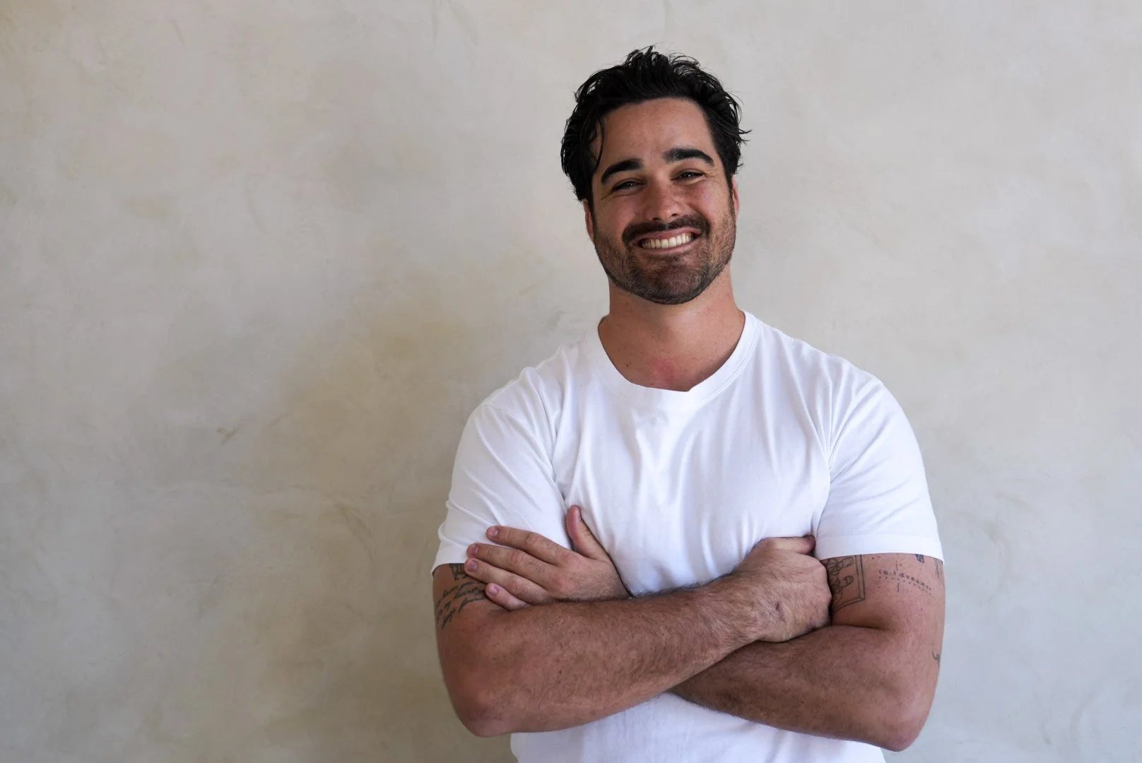 A man with dark hair and a beard smiling with arms crossed, standing against a light-colored wall, wearing a white t-shirt.