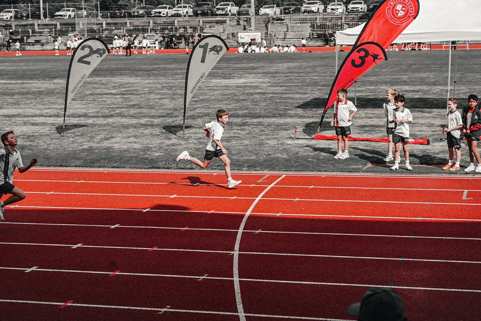 Children running on a red track during a race, with some standing near flags marked 1, 2, and 3, at an outdoor sports event.