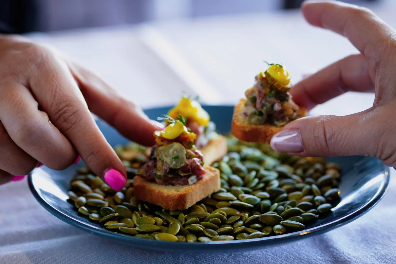 Two hands with manicured nails, one with pink nail polish and the other with light purple, are placing small slices of toasted bread topped with a colorful spread and garnishes onto a blue plate filled with pumpkin seeds.