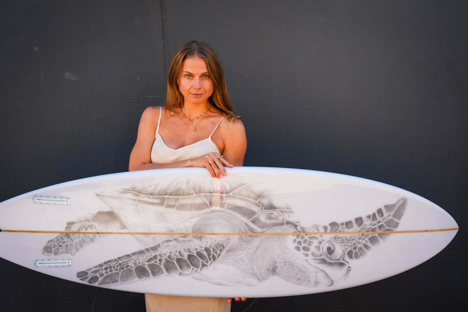 Woman with long brown hair in a white tank top holding a surfboard with a turtle illustration against a black background.