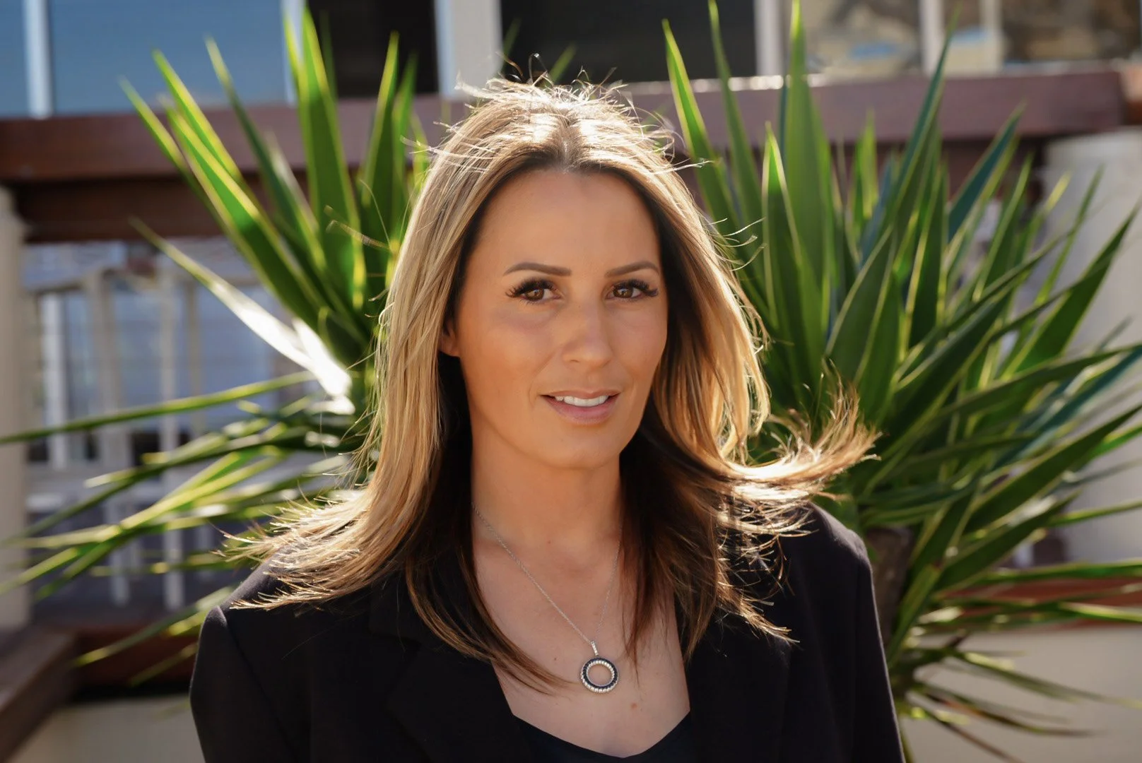 A woman with light brown hair and a black blazer standing outdoors in front of a large green plant, smiling at the camera.