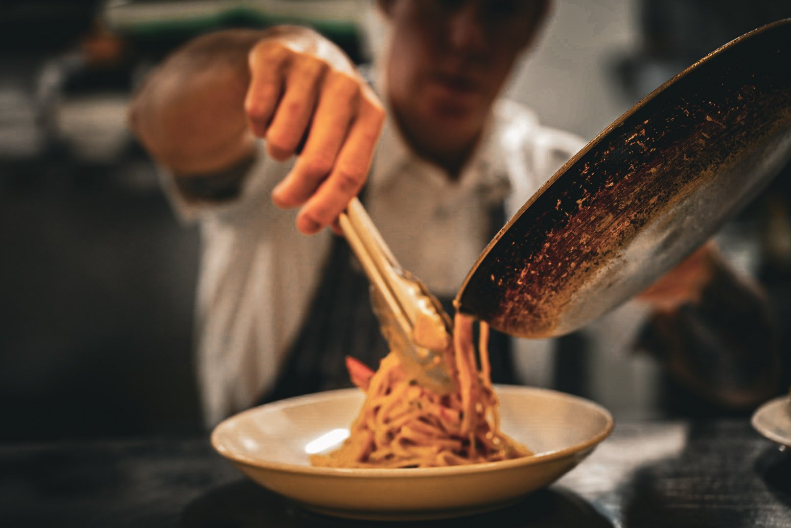 Chef serving noodles onto a plate in a restaurant kitchen.