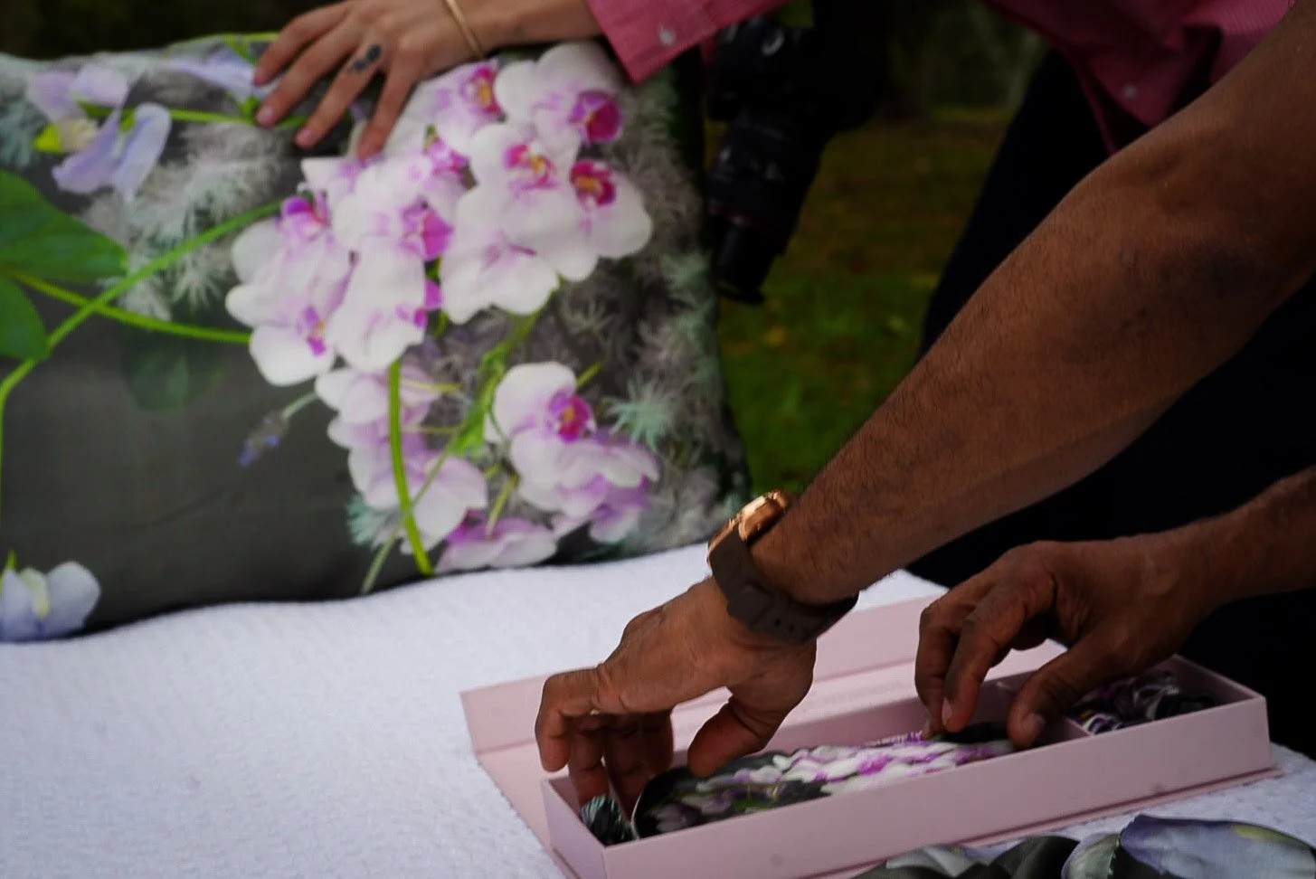 Person arranging a floral arrangement of pink and white orchids on a table outdoors, with a pink box and a backdrop of greenery.