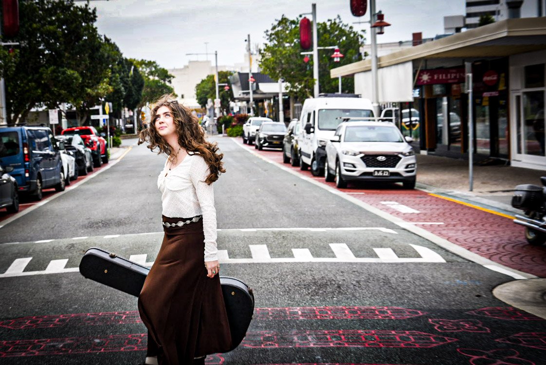 A young woman with curly hair holding a guitar case stands on a city street crosswalk, looking up. There are parked cars and shops lining the street.