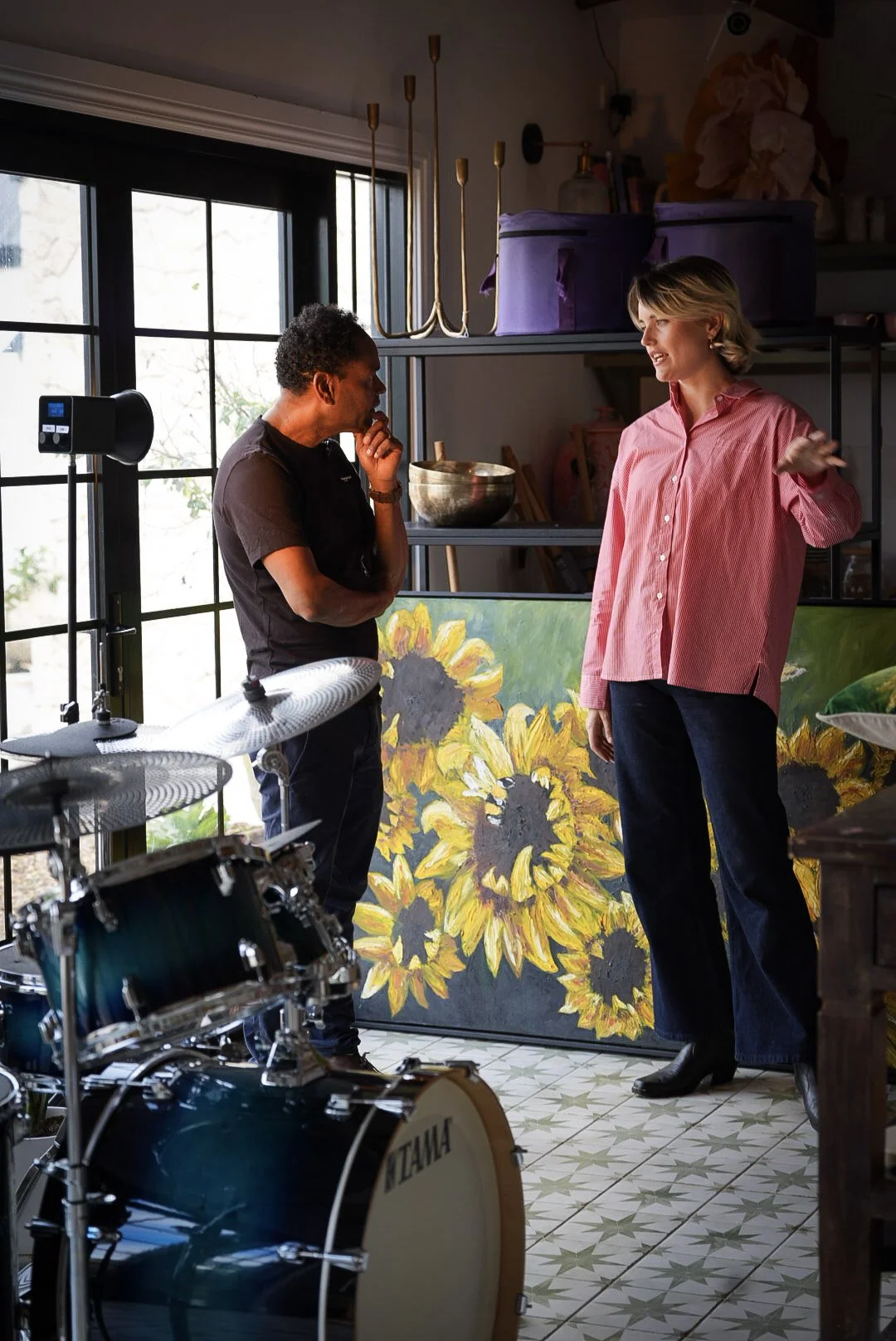 Two women having a conversation in an art studio, with sunflowers painted on a canvas behind them, a drum set in the foreground, and large windows letting in natural light.