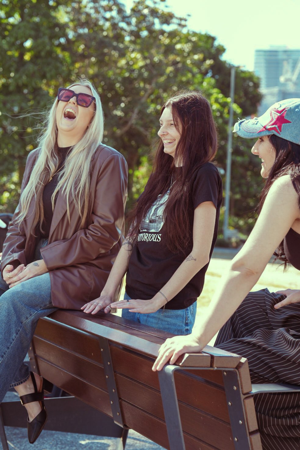 Three women sitting on a park bench outdoors, laughing and chatting, with trees and buildings in the background.