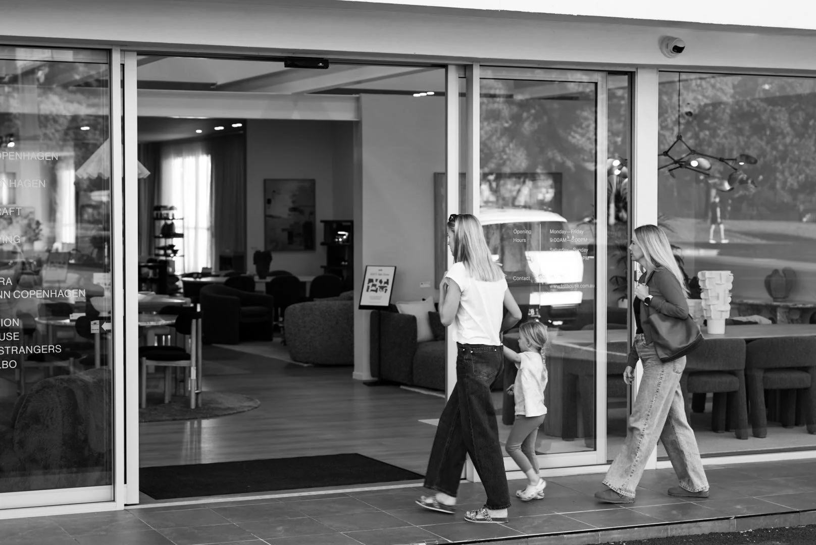 Three women and a child walking past a cafe with large glass windows. Inside the cafe, there are seating areas with chairs and sofas, as well as decorative items and lighting fixtures.
