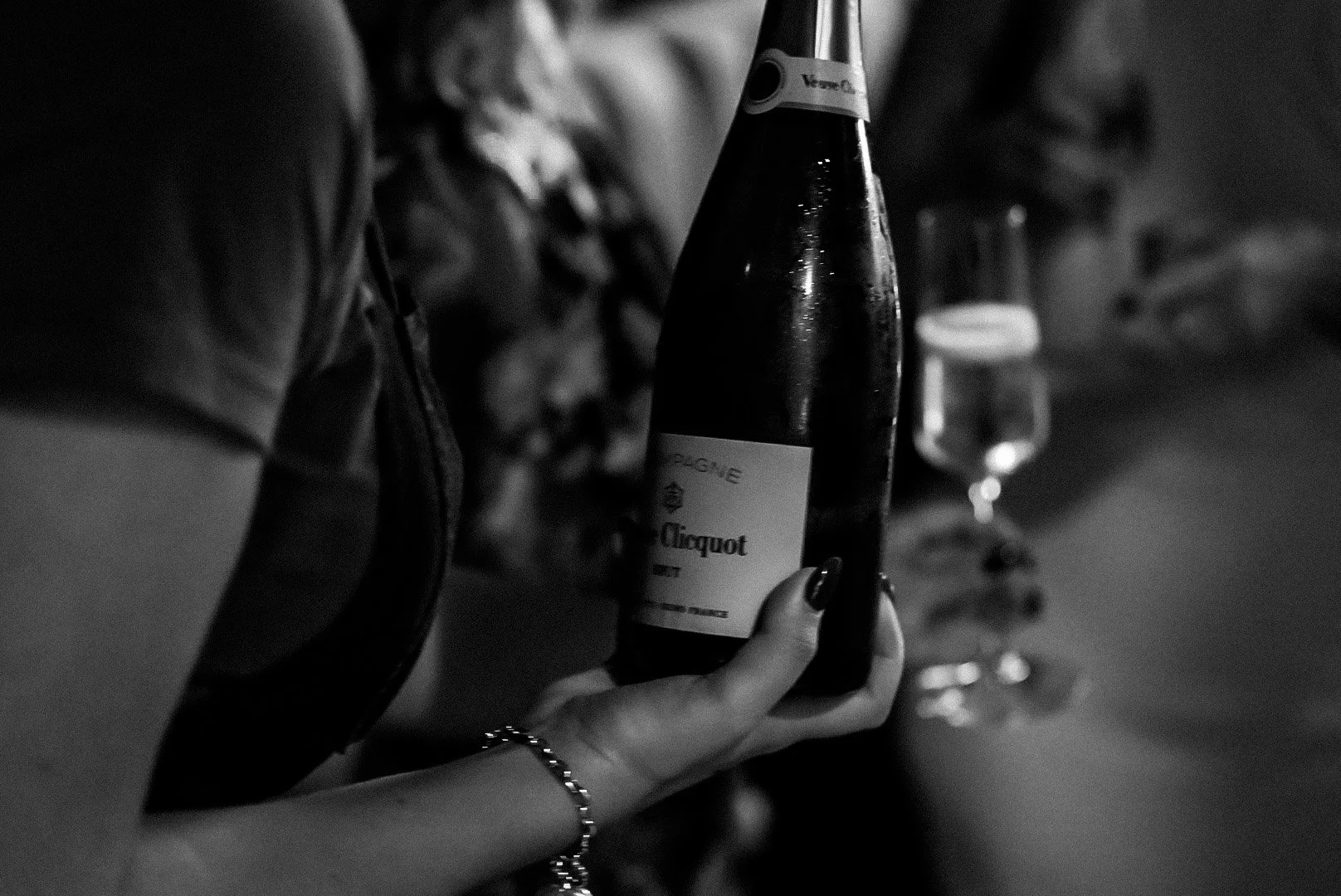 Person holding a bottle of champagne with a label that reads 'Moet & Chandon' at a bar with other drinks in the background in black and white.