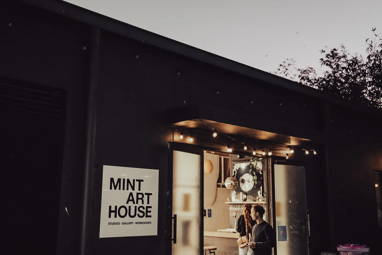 Entrance to Mint Art House, a studio, gallery, and workshop space, with people inside and an illuminated sign on the left side.