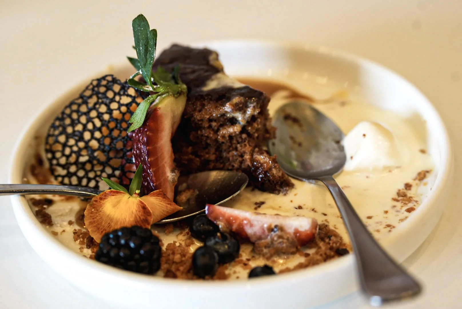 Close-up of a dessert bowl with a slice of chocolate cake, fresh strawberries, blackberries, and edible flowers, topped with whipped cream and chocolate crumbles, with spoons in the dish.