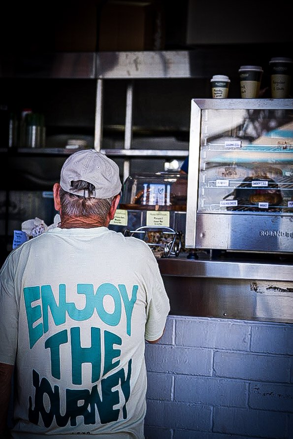 A man with gray hair wearing a light-colored shirt with the phrase 'Enjoy the Moment' on the back, facing away while standing at a counter in a cafe or food shop.