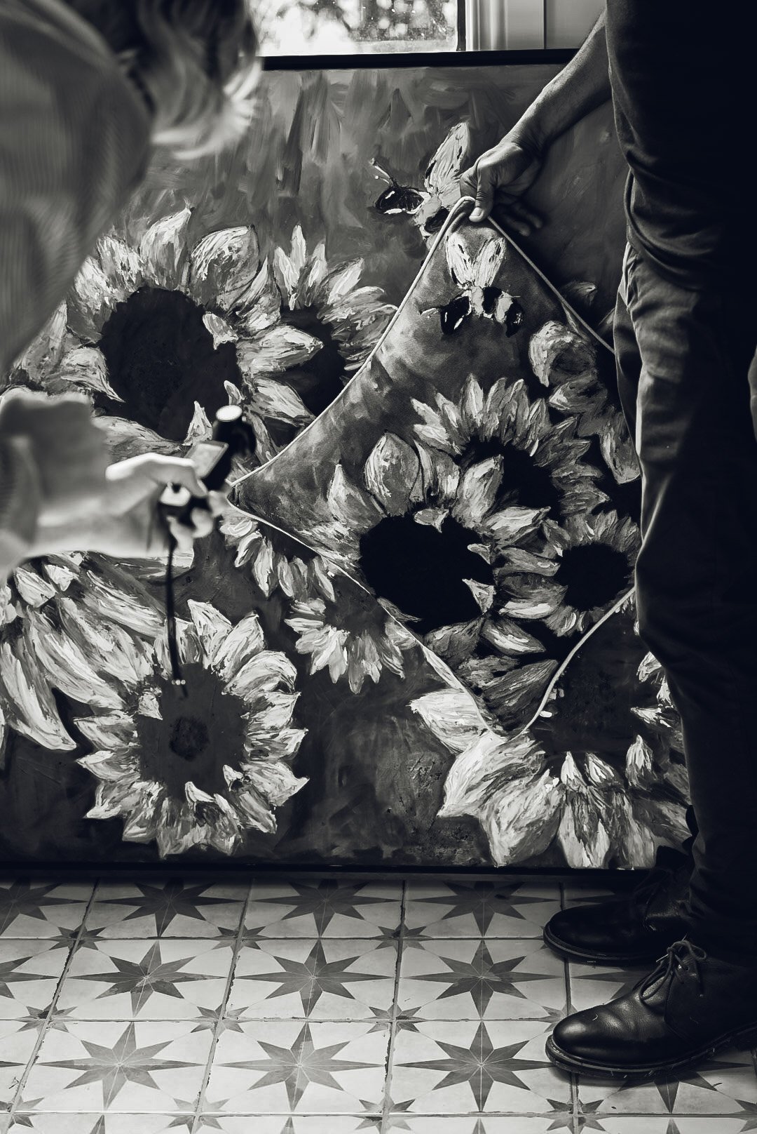 Two people setting up a sunflower painting with a black and white abstract background, one holding a painting and the other adjusting it, standing on patterned floor tiles.