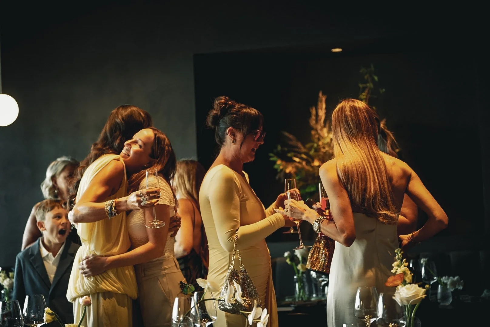 Group of women and children at a celebration, holding champagne glasses, in a dimly lit room with floral centerpieces.
