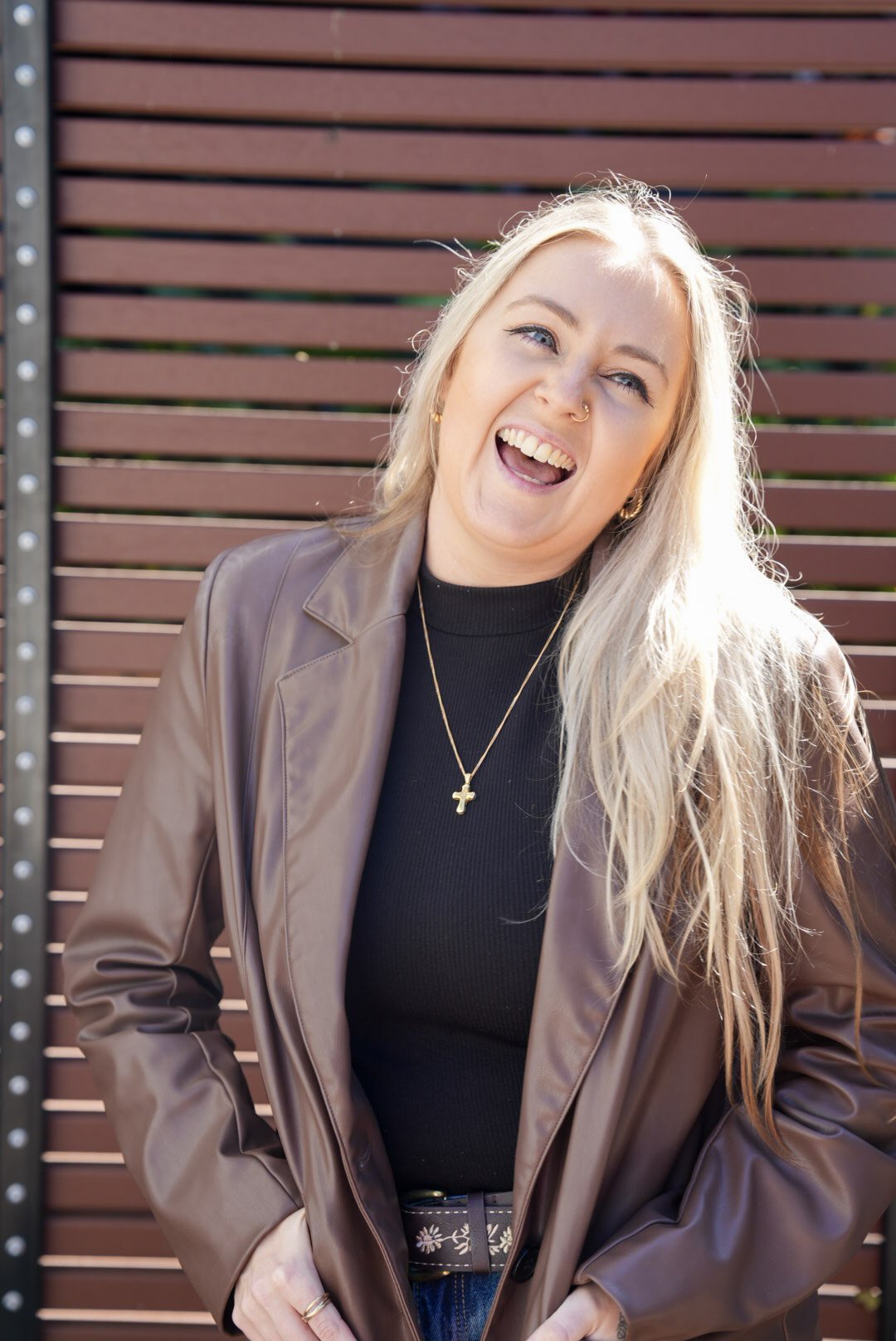 A woman with long blonde hair smiling and sticking her tongue out, standing in front of a brown wooden slatted fence.