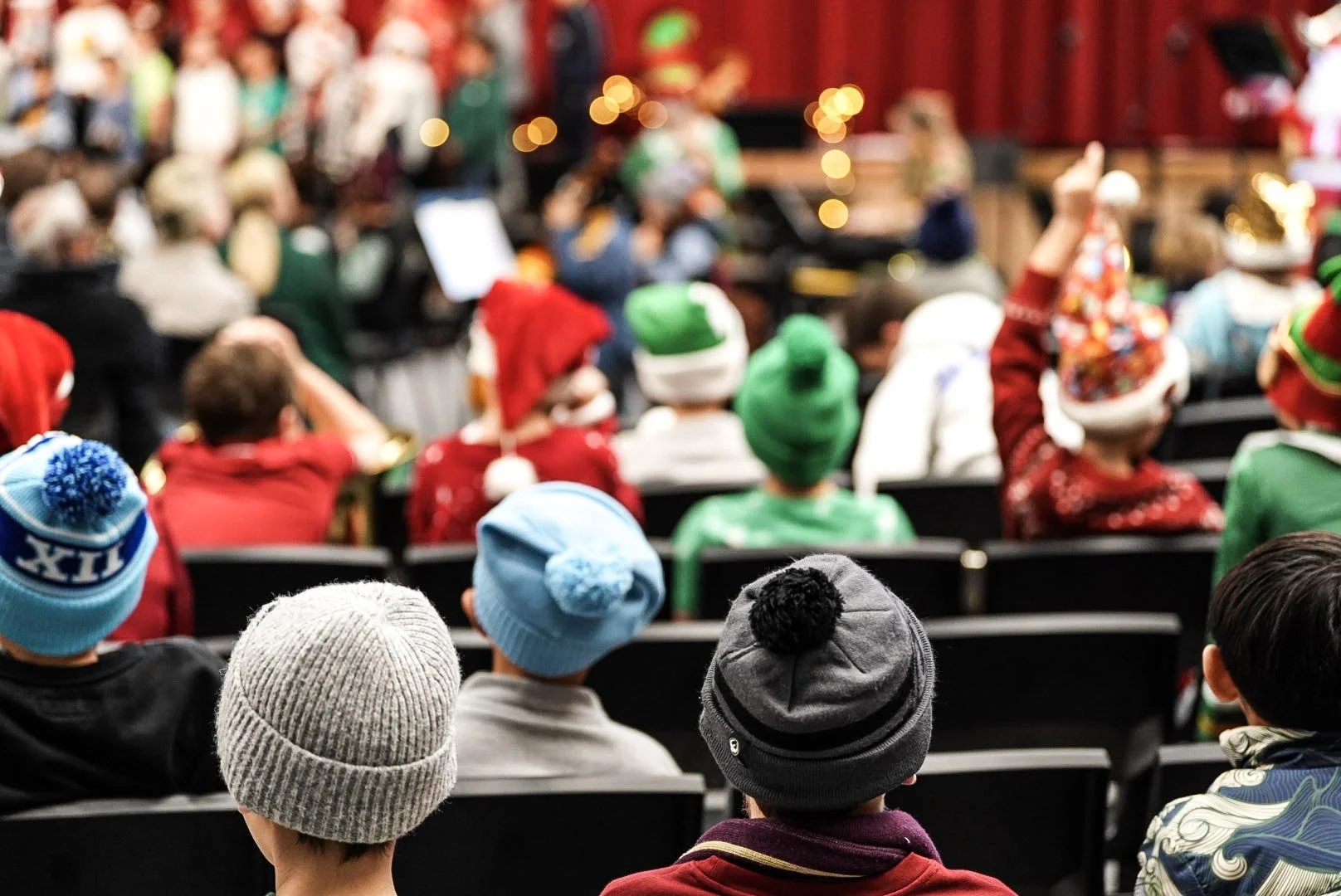 Children wearing colorful holiday hats and sweaters sitting in a theater or auditorium, facing a stage with a Christmas performance or event, with a blurred background of more children and stage lights.