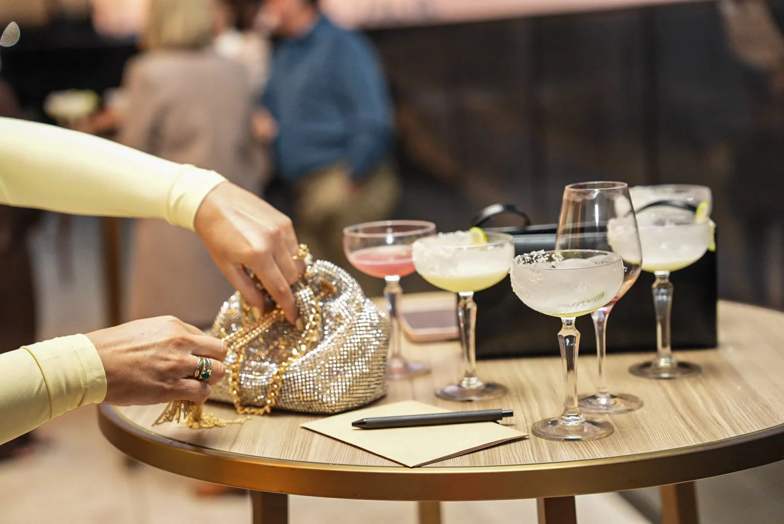A person in a yellow long-sleeve top is reaching for a sparkly, beaded clutch bag with gold chain strap on a round wooden table. The table also has several cocktail glasses containing various drinks, a black purse, a notepad, and a pen.