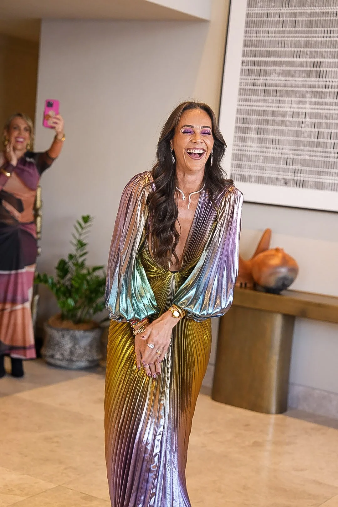 Woman in a shimmering, multicolored dress laughing, with another woman taking a photo of her in the background.