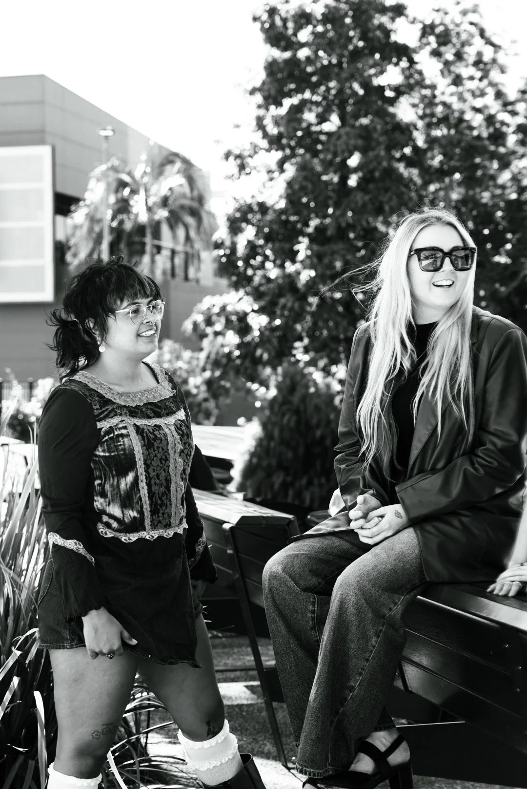 Two women, one standing and one sitting on a park bench, engaging in conversation outdoors with trees and buildings in the background in black and white.