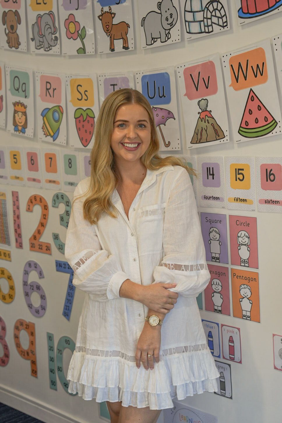 A woman standing in front of a colorful educational wall with alphabet letters, numbers, and illustrations for children, smiling at the camera.