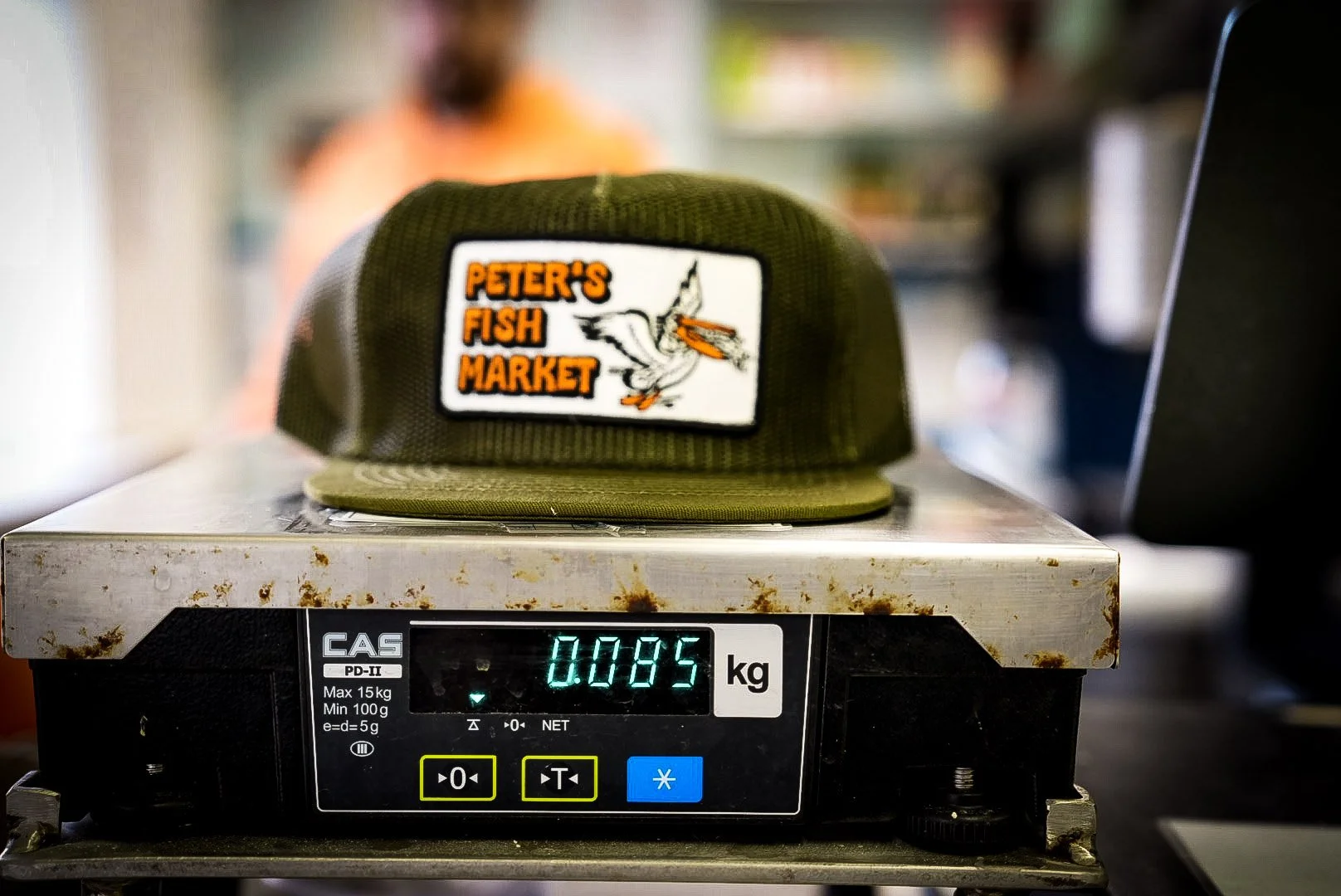 A cap with the logo of Peter's Fish Market placed on a metal scale showing a weight of 0.085 kilograms.
