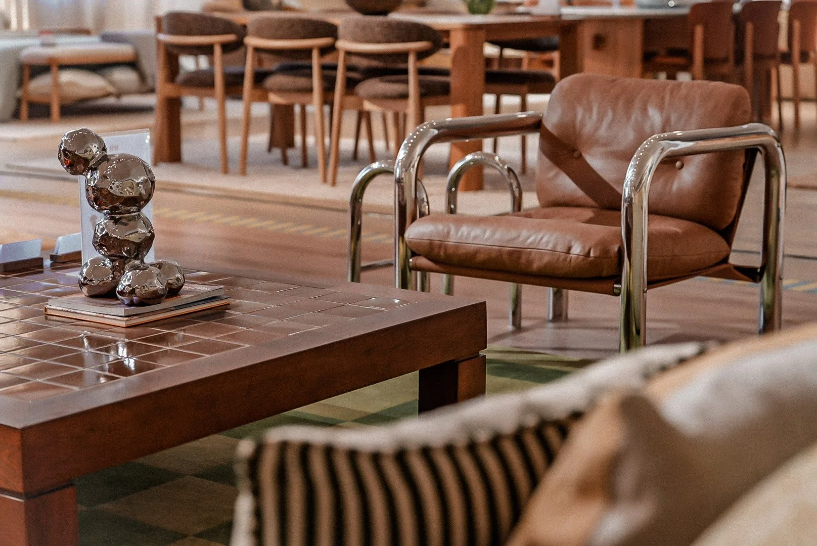 Interior of a living room featuring a brown leather chair with chrome arms, a wooden table with a decorative silver sculpture, and various dining chairs in the background.