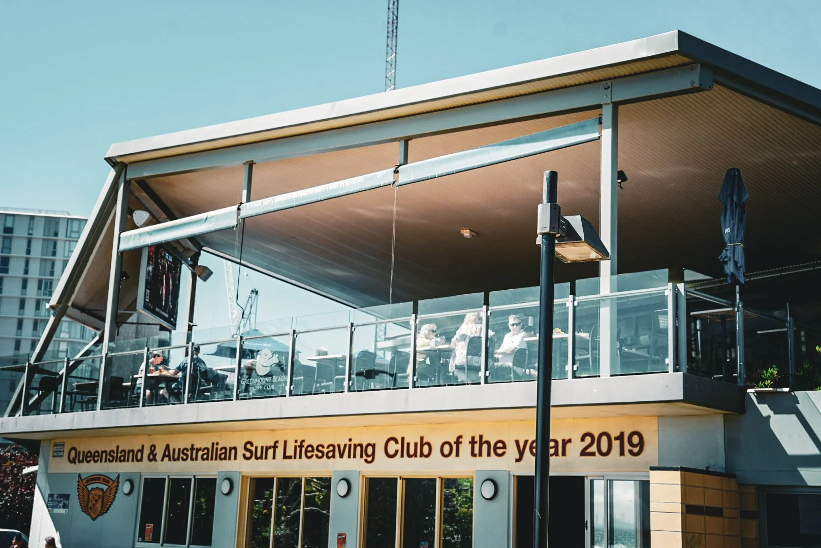 Two-story beach club with people sitting at tables on the upper deck, glass railing, and signage indicating the Queensland & Australian Surf Lifesaving Club of the year 2019. City buildings and a clear sky in the background.