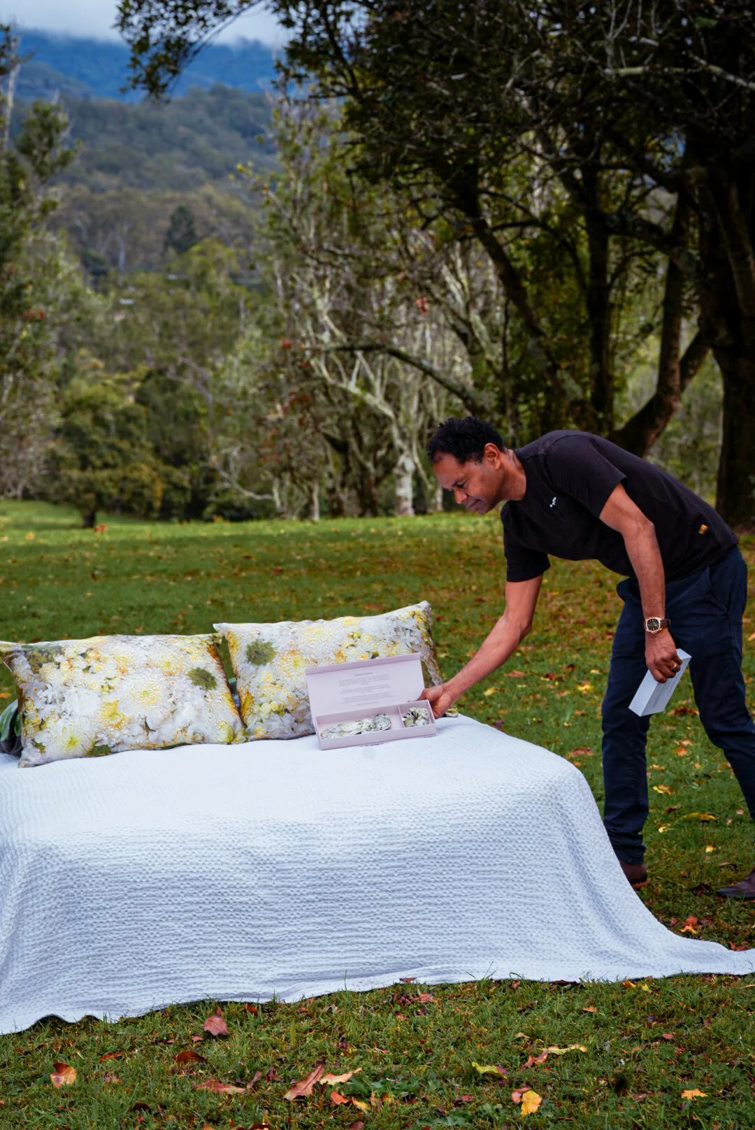 A man is crouching next to an outdoor bed with white and floral pillows, holding a box and a booklet, in a park with trees and mountains in the background.