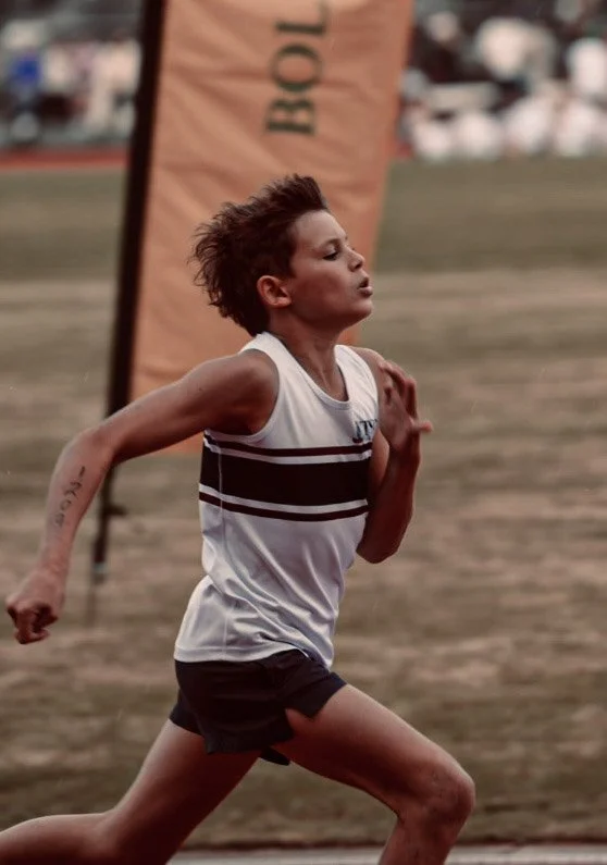 A female athlete running on a track during a race, with a banner behind her and spectators in the background.