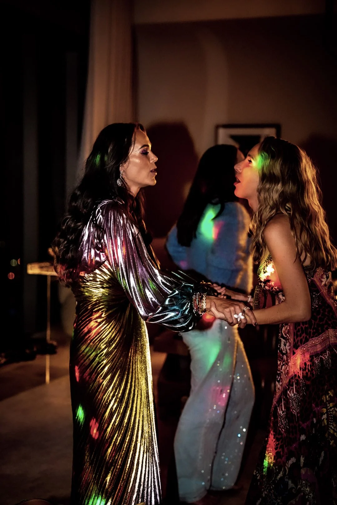 Two women shaking hands at a party or dance event, with colorful lights. One woman has long dark hair and wears a shiny, metallic dress; the other has long wavy hair and wears a patterned dress.